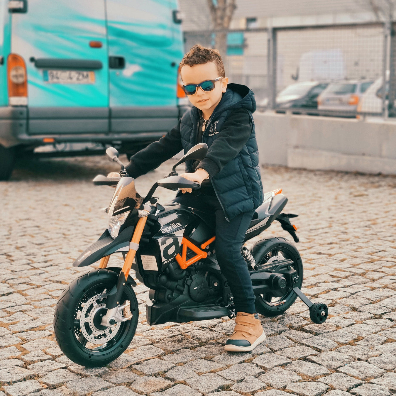 Een jongen met een zonnebril zit stoer op een elektrische kindermotor en kijkt recht in de camera, terwijl hij op straat stilstaat.